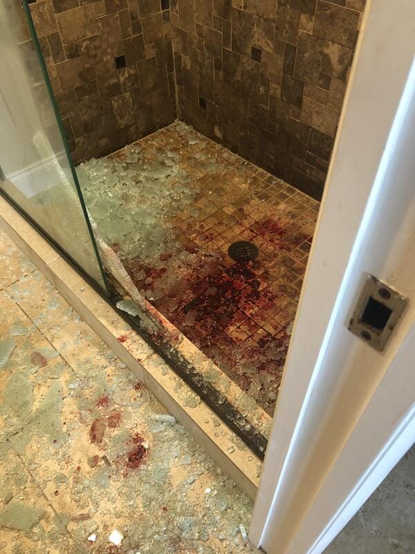 lood-stained tiled shower floor with shattered glass scattered outside, indicating a biohazard scene requiring professional blood cleanup and trauma restoration services