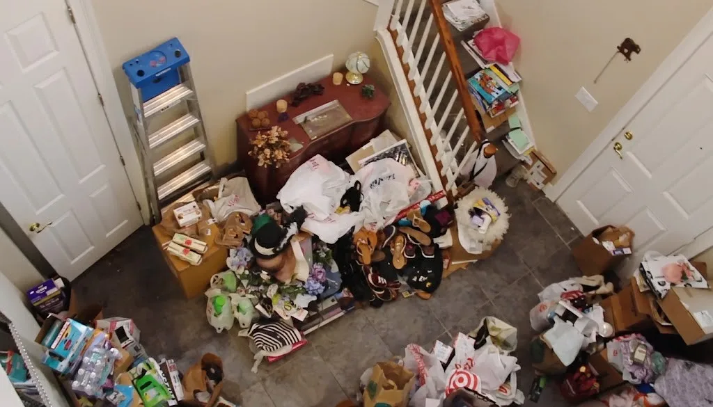 An overhead view of a heavily cluttered home entryway and staircase filled with piles of bags, boxes, shoes, and household items.