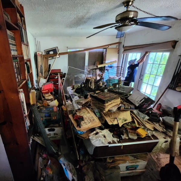 Cluttered room packed with stacks of old newspapers, tools, boxes, and debris covering a table and floor, illustrating severe hoarding conditions; worker opening window blinds to let in light during professional spring cleaning and decontamination service by Spaulding Decon, highlighting deep cleaning, junk removal, and property restoration.